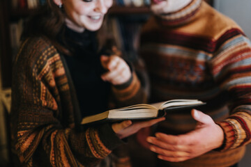 Two close friends enjoy reading a book together while wearing warm, patterned sweaters inside.