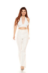 Fototapeta premium Confident woman in all-white halter crop top and wide-leg pants, smiling and walking with studio lighting, embodying modern fashion elegance
