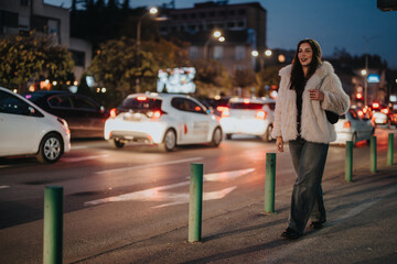 A woman wearing a fashionable coat walks on a city street at dusk, surrounded by car traffic and streetlights. The urban ambiance is lively, depicting a moment of elegance within a vibrant setting.