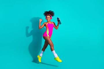 Young athletic woman in vibrant sportswear holding a water bottle on a bright teal background,...