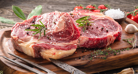 Raw beef steaks with herbs and spices ready for cooking are on wooden board.