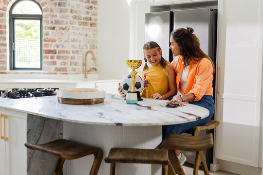 Mother and daughter celebrating soccer trophy in modern kitchen, smiling together