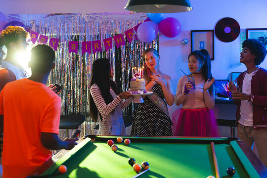 Diverse Teenage friends celebrating birthday with cake and drinks around pool table at party, at hom