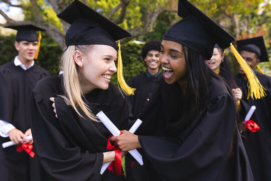 Graduating diverse students smiling and embracing while celebrating with diplomas in garden