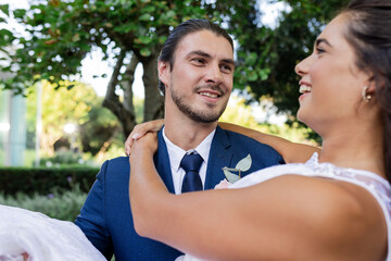 Groom in blue suit joyfully carrying bride outdoors, both smiling and celebrating