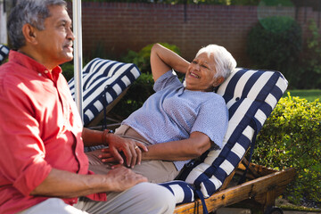 Senior diverse couple relaxing on patio chairs, enjoying conversation in sunny backyard