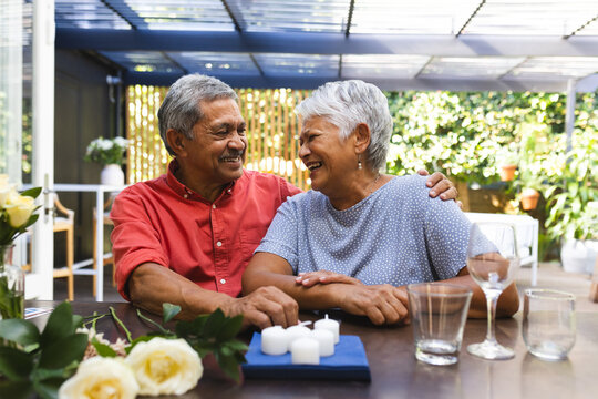 Elderly diverse couple sitting at table, smiling and enjoying time together in garden