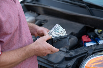 Man holds dollar bills while assessing car repairs with determination. A man examines his vehicle's...