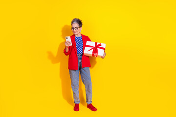 Stylish senior woman holding a gift box and smartphone on a vibrant yellow background, showcasing casual fashion