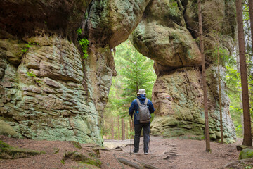 Man holding hiking sticks walks through natural stone gate called Wrota in Skalne Grzyby rocks in Stolowe Mountains National Park, Poland