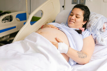 Pregnant Latina Colombian woman lying on a hospital bed, smiling and caressing her belly. In a maternity clinic, waiting to give birth with happiness, tenderness, and well-being