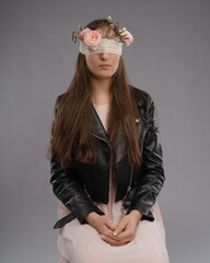 A girl with closed eyes, adorned with a blindfold decorated with flowers and branches, wearing a leather jacket. A conceptual portrait with elements of symbolism, mystery, and natural aesthetics.