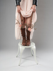 A conceptual shot of a girl standing on a white chair, wearing a flowing pastel dress and a leather jacket. Her boots are decorated with flowers and branches, creating an artistic and symbolic image