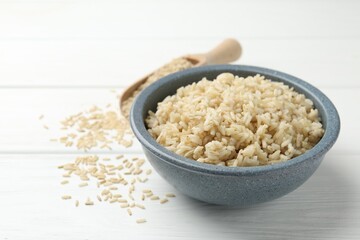 Tasty brown rice in bowl and raw grains on white wooden table, closeup
