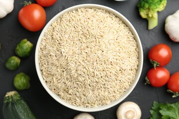 Raw brown rice in bowl among fresh products on dark textured table, flat lay