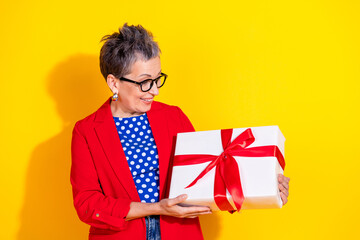 Charming middle-aged woman holding gift box against vibrant yellow background, combining stylish...