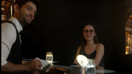 Focusing on beautiful woman guest sitting in special drink gin fizz at luxurious counter bar at vibrant nightlife party atmosphere. Smart bartender wiping glass while talking with customer. Vinosity.