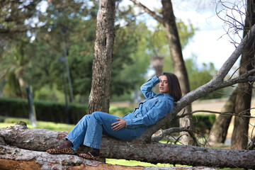 Young, beautiful, brunette, Hispanic woman, wearing a denim suit, relaxed and calm looking at the camera, lying on the fallen trunk of a tree, in the nature, receiving the sun rays.