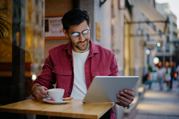Obraz premium Male freelancer enjoys coffee while working on a tablet at an outdoor café in the evening. Focused on the screen, man manages tasks remotely, combining work and leisure in a stylish urban setting