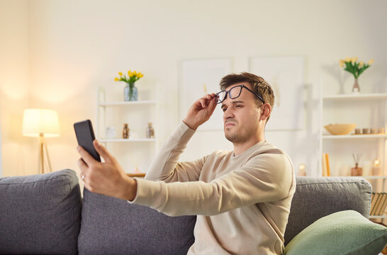 Unhappy and upset man spending leisure time online in internet with mobile phone at home, struggling to see due to bad eyesight. Challenges of using technology while dealing with vision issues.
