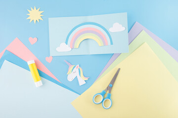 handmade rainbow card with white clouds and pink hearts. Surrounding elements include unicorn cutout, sun, scissors, glue stick, and paper sheets. Useful for craft stores, schools, and DIY blogs