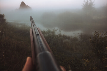 Hunting shotgun barrel aiming during foggy morning on river bank during hunting season as hunting background in old vintage style 