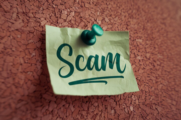 Green note pinned to brown corkboard, displaying the word "Scam" in a stylized script font, representing warning, fraud, and online security.