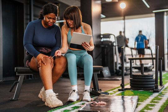 Overweight woman is sitting on a bench in a gym, pointing at her knee while discussing her pain with her personal trainer, who is holding a tablet and listening attentively