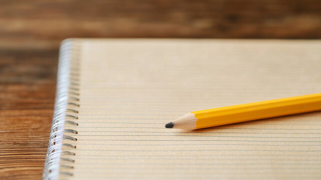A close-up of a yellow pencil resting on a spiral notebook with lined pages, set against a rustic wooden background.