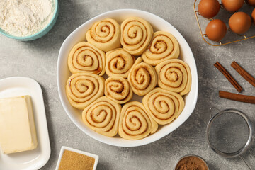 Raw cinnamon rolls in baking dish and different ingredients on grey table, flat lay