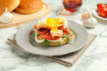 Tasty brunch. Poached egg, ham, tomato, arugula and bun on white marble table, closeup