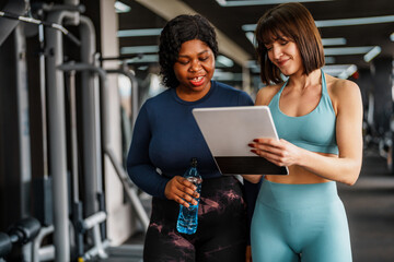Smiling personal trainer demonstrating a training plan on a digital tablet while an overweight woman holds a water bottle, both engaged in a supportive discussion inside a gym