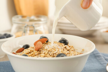 Woman pouring milk into bowl with oatmeal, blueberries and almonds at table, closeup
