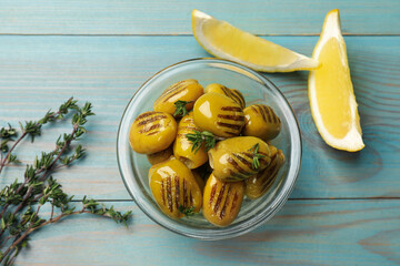 Grilled green olives in bowl, thyme and lemon wedges on light blue wooden table, flat lay