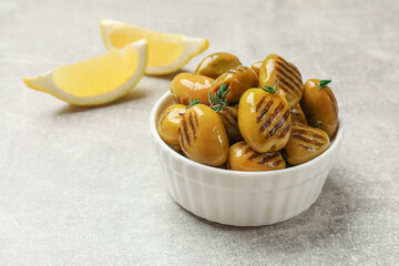 Grilled green olives with thyme in bowl and lemon wedges on gray textured table, closeup. Space for text