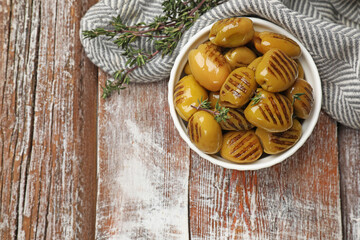 Grilled green olives with thyme in bowl on wooden table, flat lay. Space for text