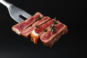 Carving fork with pieces of delicious beef meat and thyme on black background, closeup