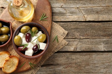 Marinated olives with feta cheese, bread pieces, oil and rosemary on wooden table, top view. Space for text