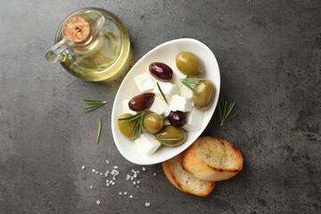 Marinated olives with feta cheese, bread pieces, oil, salt and rosemary on grey table, flat lay