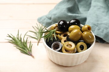 Different delicious marinated olive rings, whole ones and rosemary in bowl on light wooden table, closeup