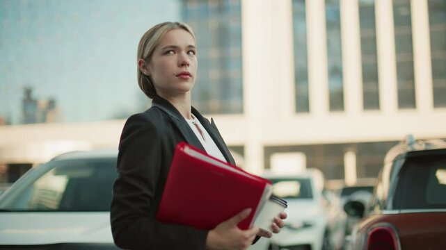 Side view of remote worker with hair clip holding red folder and coffee cup glancing left with parked cars and modern office building background during workday in bright urban setting