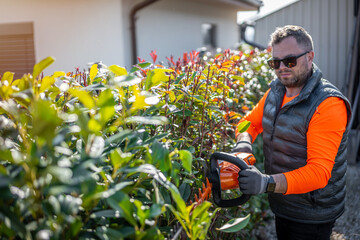 Man dressed in reflective workwear holding hedge trimmer in his hands. Bush pruning work. Gardening...