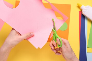 Woman cutting pink paper on yellow background, top view. Child handmade craft