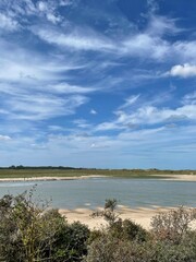 Serene coastal landscape featuring a calm body of water bordered by sandy shores and lush vegetation in the foreground. The sky dominates the scene, painted with soft, wispy white clouds.