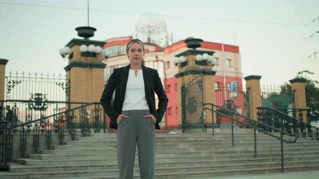 Young woman confidently poses with hands on waist while standing on steps in urban area with modern buildings and metal railings behind, expressing assertiveness in professional attire