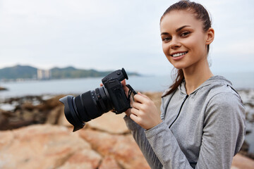 Young female photographer smiling while holding a camera outdoors by the rocky shore, showcasing a...