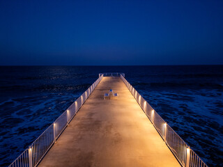 Obraz premium Long illuminated pier stretching into the dark sea at night, with calm waves and a deep blue sky, creating a peaceful and solitary coastal atmosphere