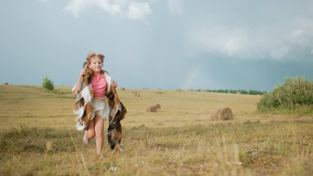 Happy little girl in pink top and shorts running across vast open field with blanket over shoulders, using it to cover head, wind blowing, golden hay bales in background