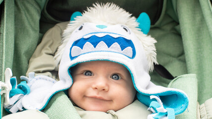 Portrait of happy smiling baby boy in white monster hat, winter clothes in green stroller. Child is 3 months old.