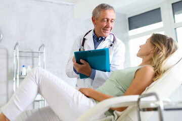 Obraz premium Friendly elderly male doctor holding document folder and engaged in conversation with young female patient lying on couch in medical office, discussing procedure details or treatment options
